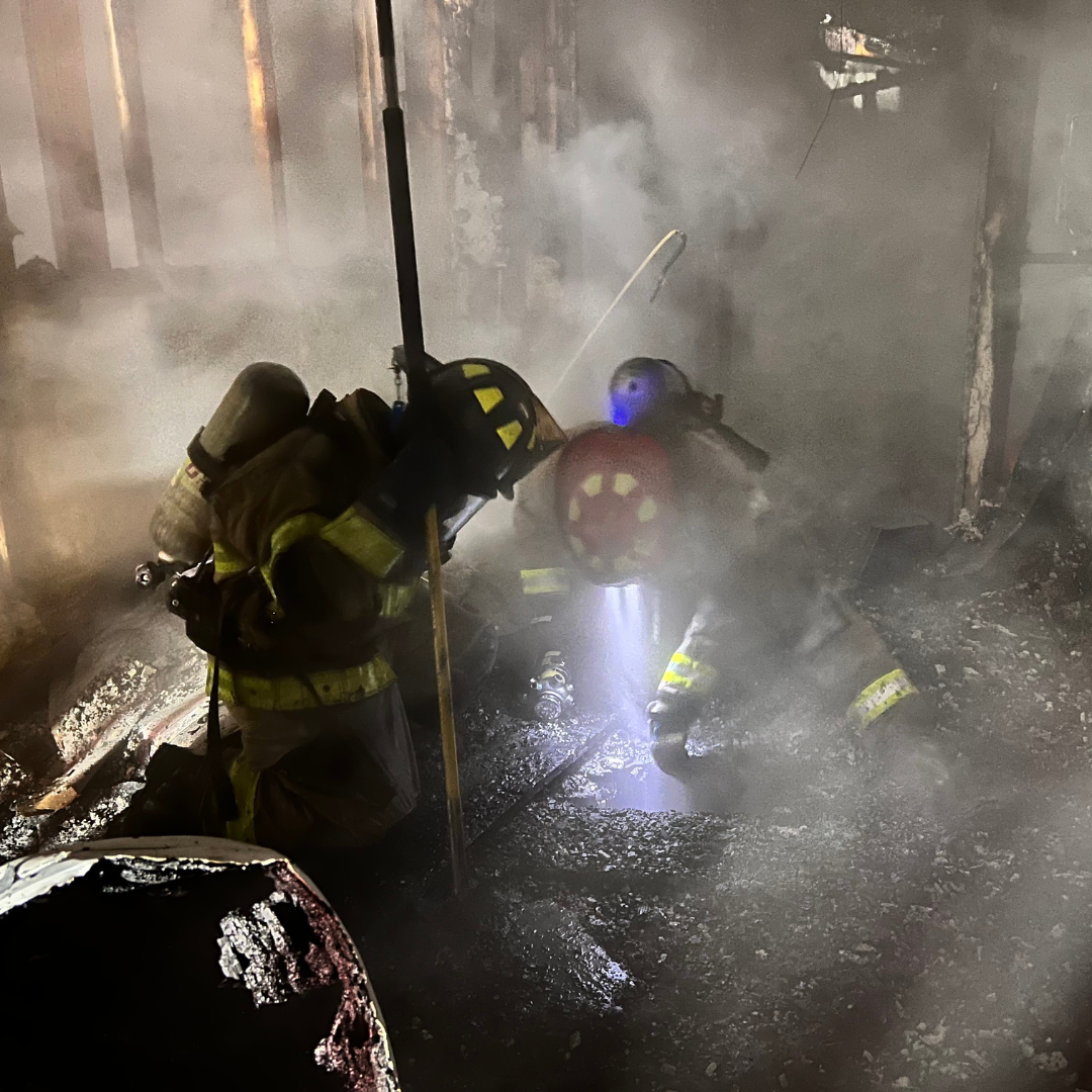 Two firefighters are in a smoky, burned down building, shining a light down a hole in the floor