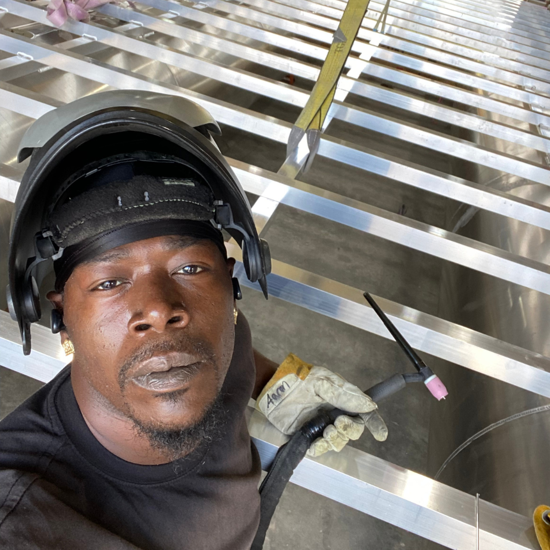 A man with a protective visor helmet on his head, holding a tool, is looking at the camera, in the background is the place he works at, Alternative Staffing