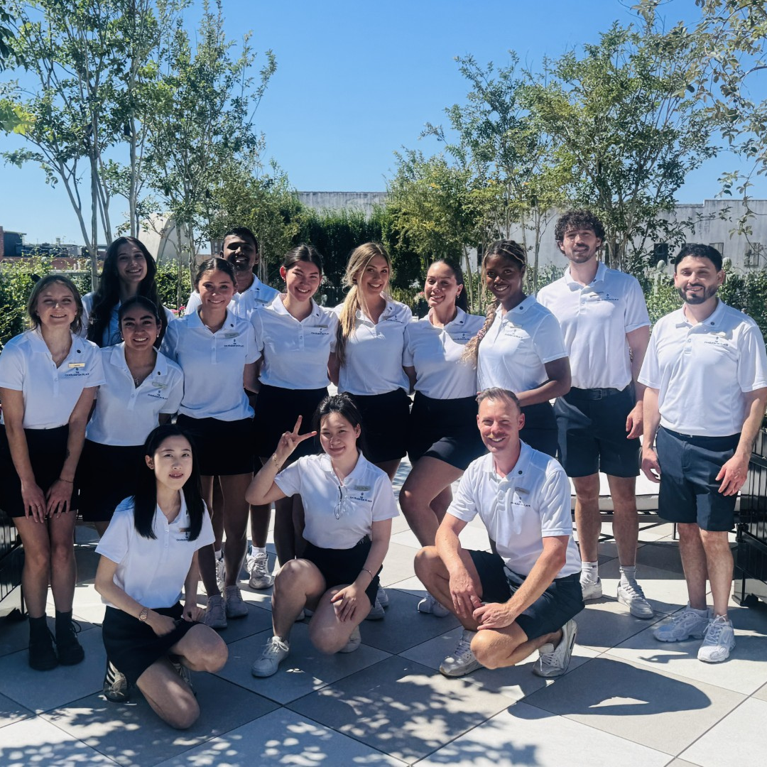 A group of workers at BHC are standing and kneeling to take a picture, all in matching white tops and blue shorts standing around trees and bushes