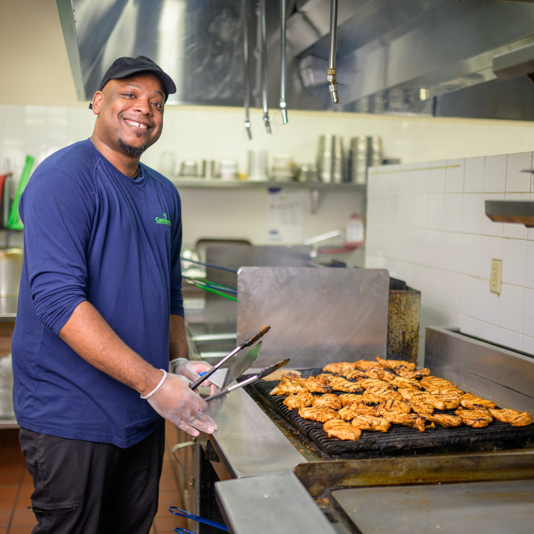 A man, holding cooking tongs, is standing in front of an indoor grill, cooking chicken, he's posing and smiling for the camera