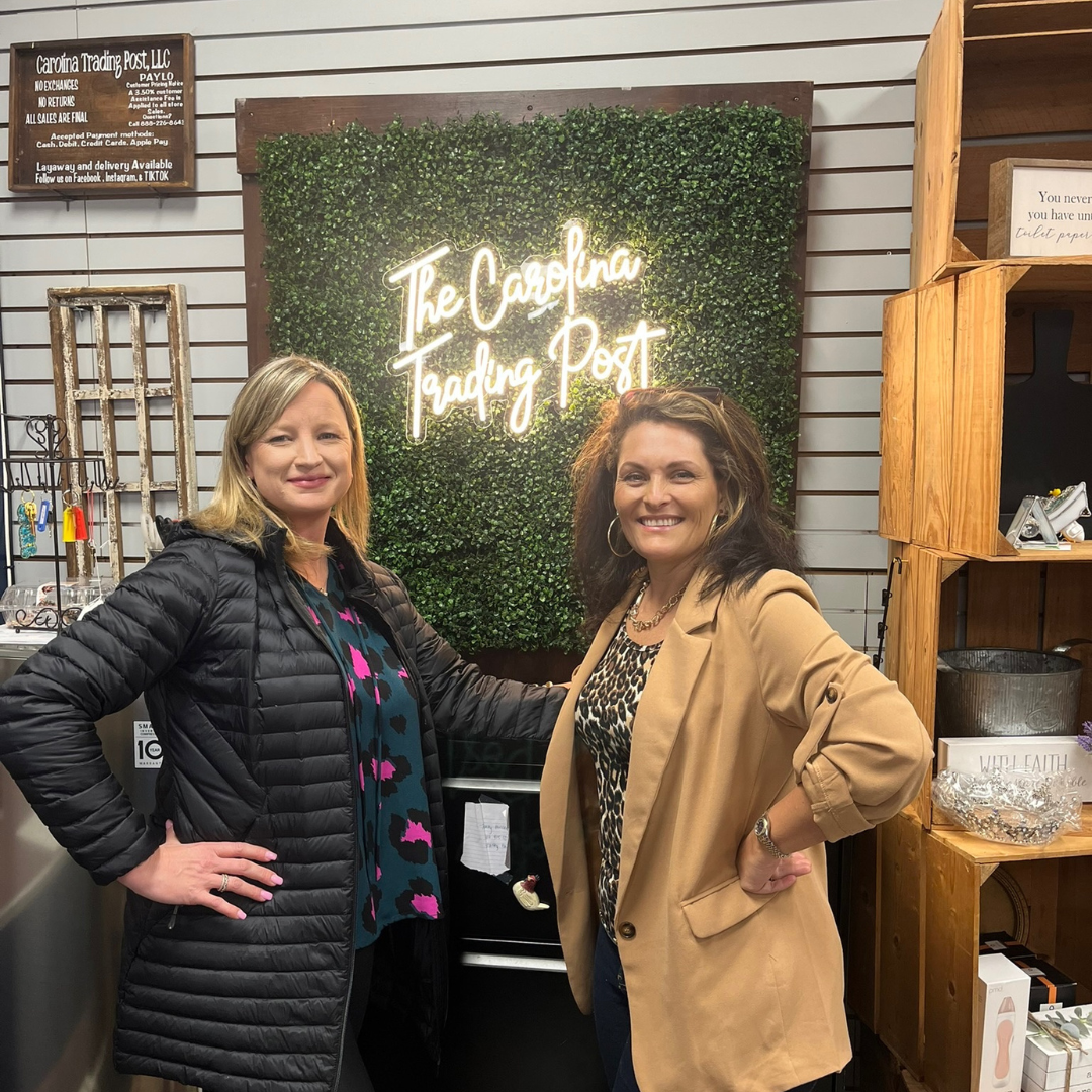 Two women, standing in front of a glowing sign that is on the wall that says "The Carolina Trading Post"
