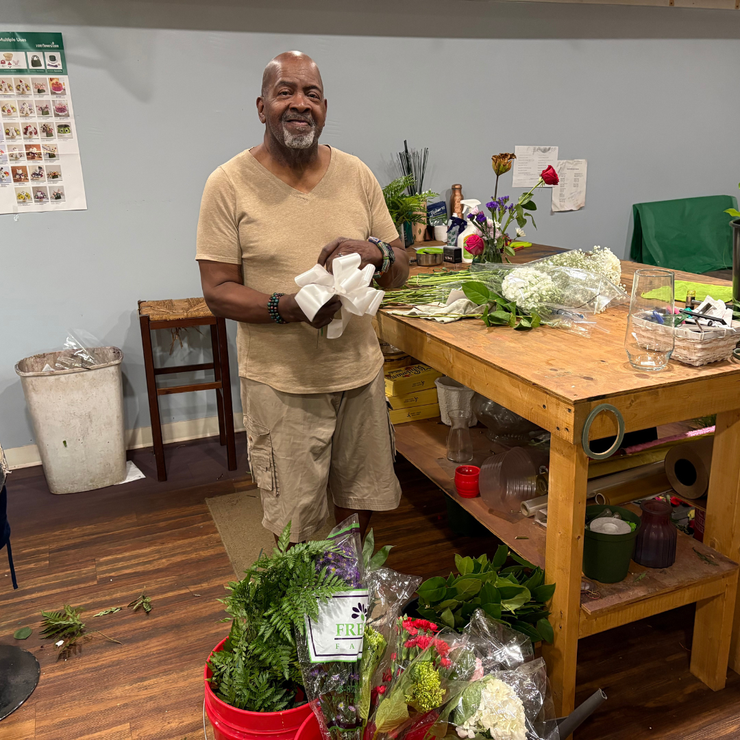 A man is standing next to a wood table, holding, and folding a satin white bow, standing in front of some plants in a bucket, about to assemble a bouquet 