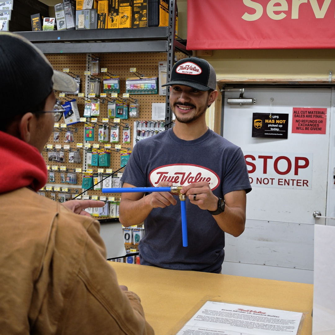 Two men, one standing behind a counter, showing a blue tool to the other man on the other side of the counter. They're having a conversation and smiling 