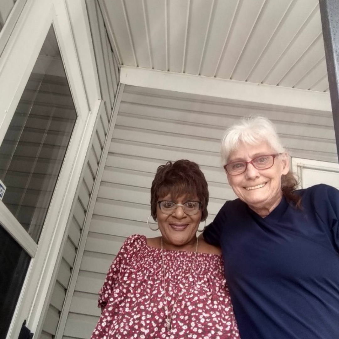 Two older ladies, are standing in front of a wall and door, arms wrapped around one another and smiling at the camera 
