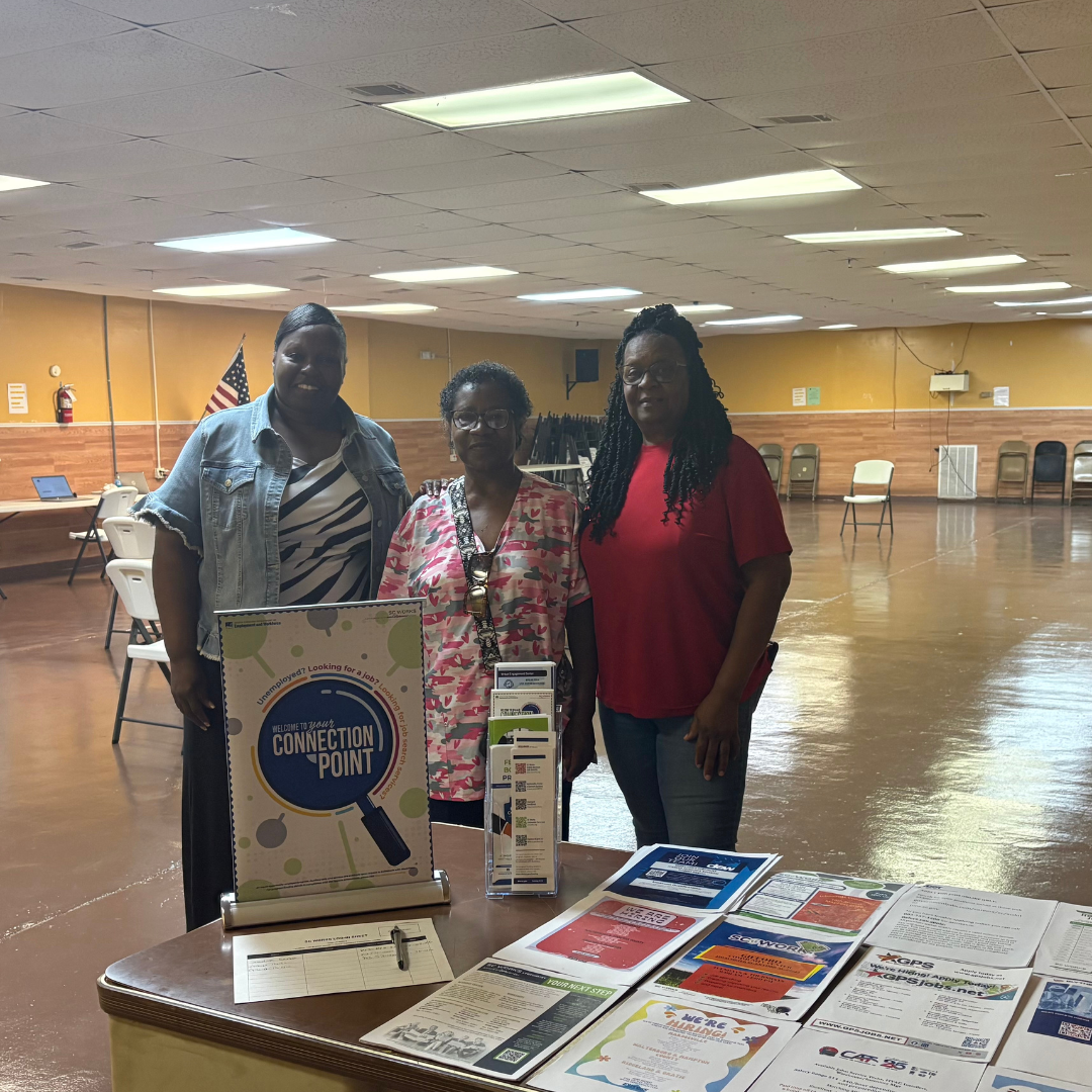 Three women are standing behind a table that have lots of flyers, brochures, and has a Connection Point flyer standing up in front of them