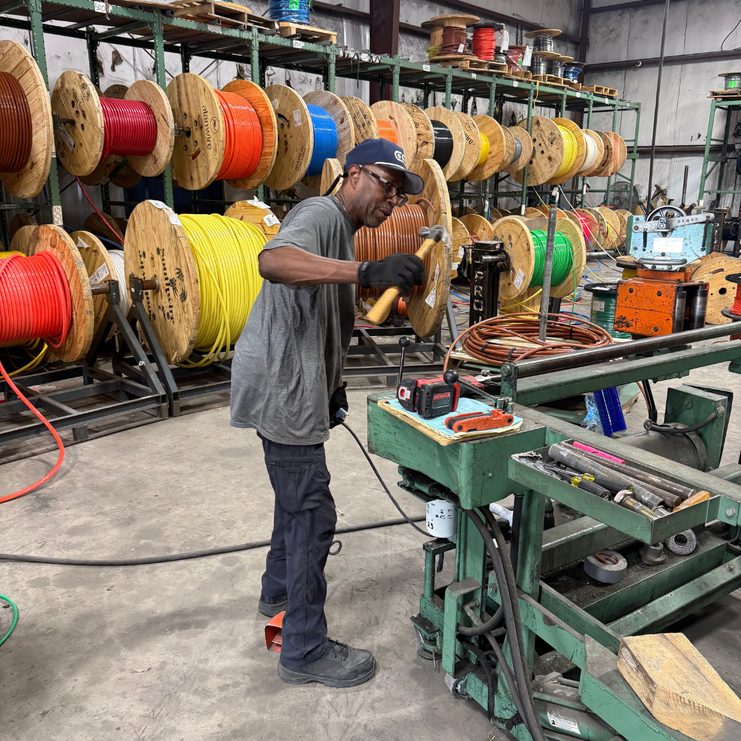 a man is leaned in front of a green table and he is holding a hammer, looking away from the camera. There are a bunch of various colored wires on wooded rolls behind him