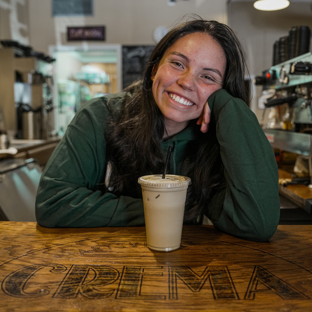 A woman is standing in front of a table, with her right arm on the table and the left elbow on the table, her hand against her face, supporting her head. She is smiling at the camera and a coffee is in front of her on the table 