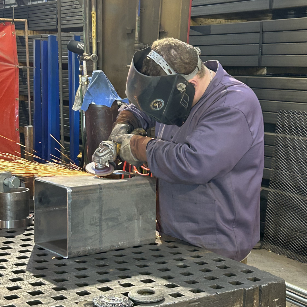 A man in protective gear and visor helmet, is using an electric tool to saw down steel, sparks flying 
