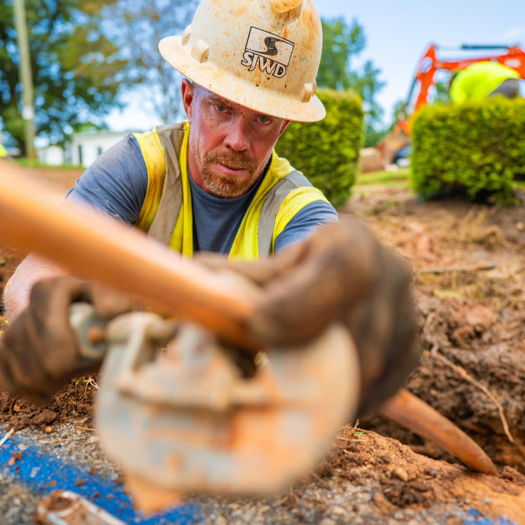 A man in a reflective vest and hard hat is using a tool to reach towards the camera, he seems to be in a hole, working on lines