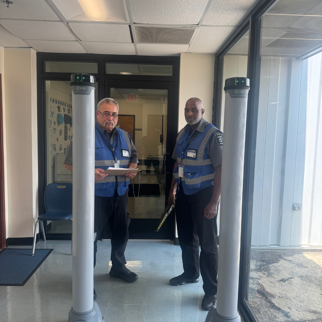 Two male security guards are standing behind a medal detector with a medal detecting wand