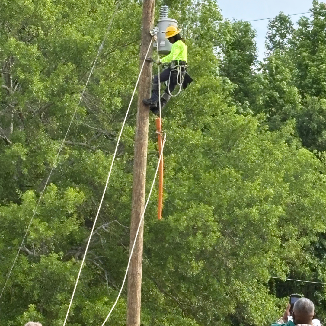 A man in a bright green shirt and hard hat is climbing a pole with wires, a lineman fixing the line