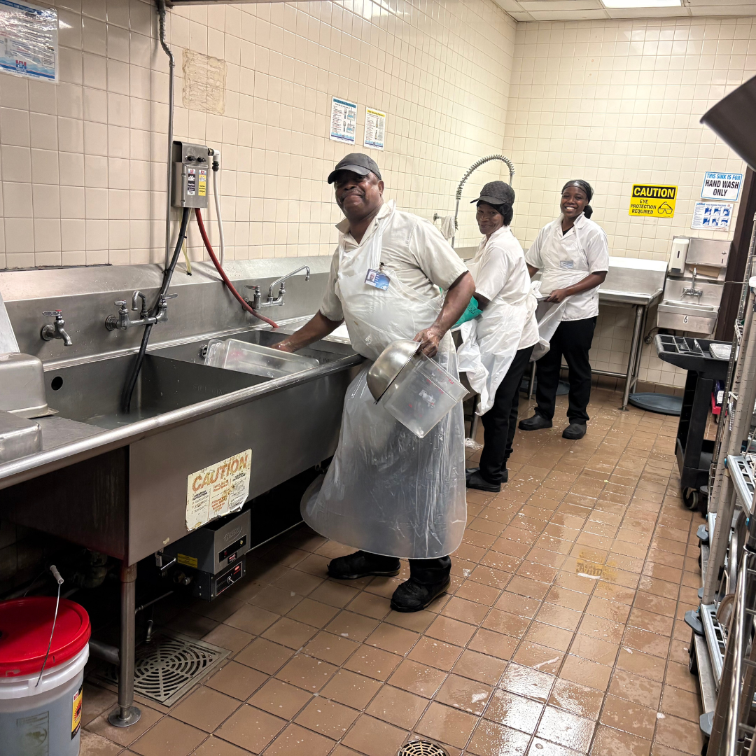 Three dishwashers are working in front of a sink while looking back at the camera and smiling 