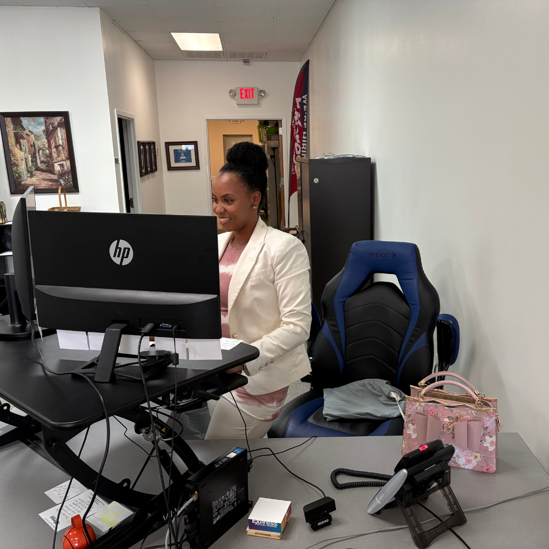 A lady is working at a desk, standing in front of two desktop computers, smiling while she does work