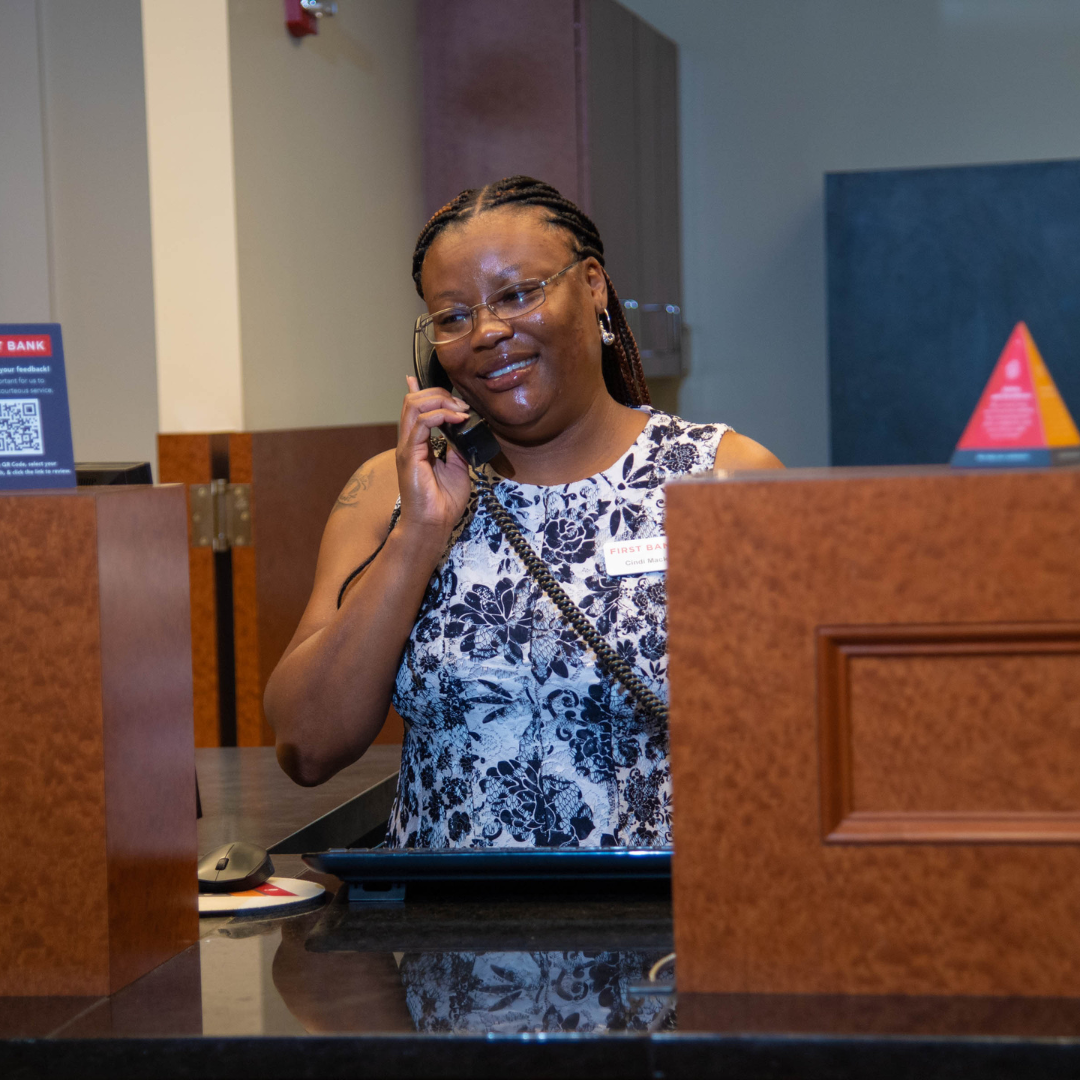 A lady behind a teller stand at a bank, is smiling, holding a phone to her ear to answer customers 
