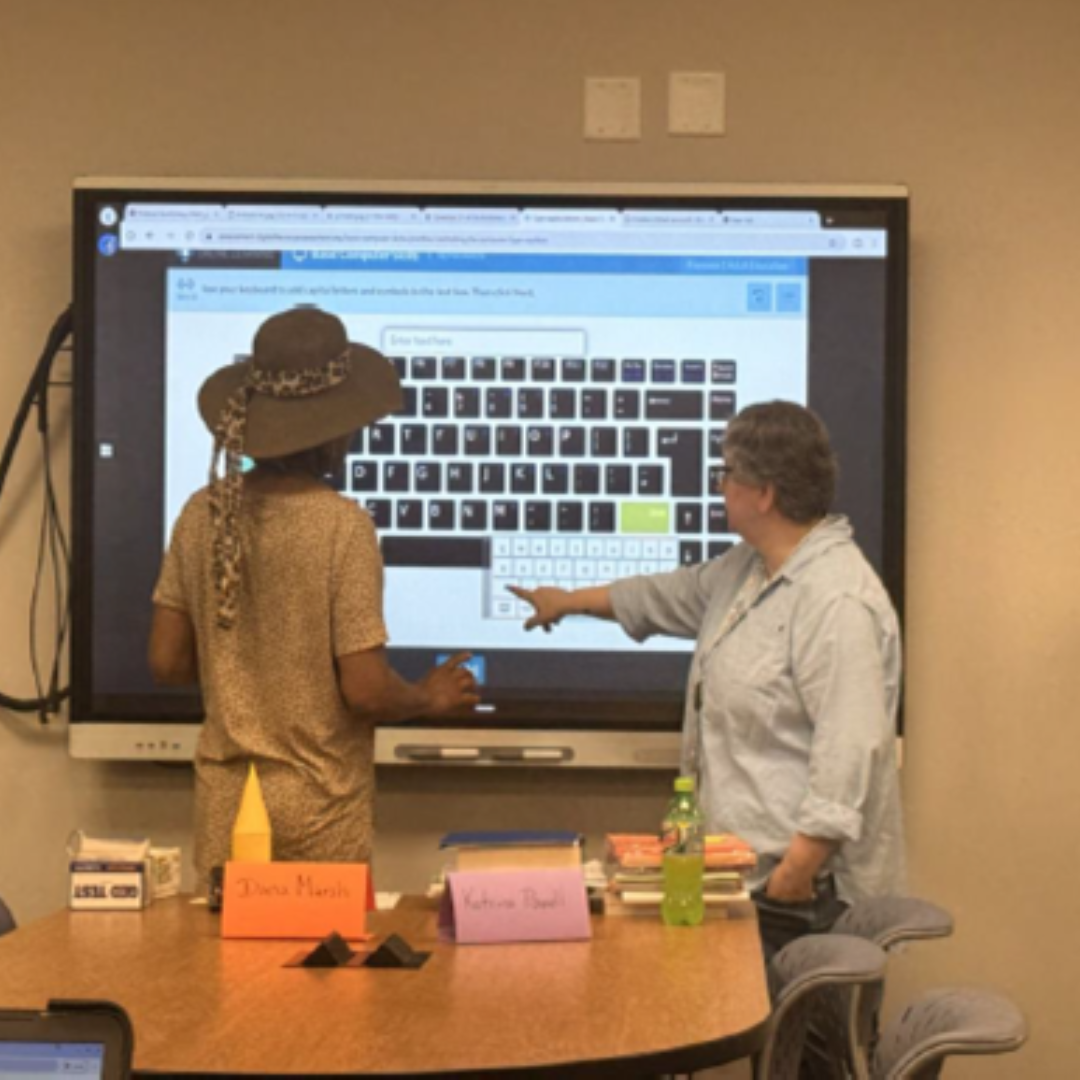 Two adults are standing up and pointing up at a smart board before them, on the wall, they appear to be pointing at a displayed keyboard on the smart board