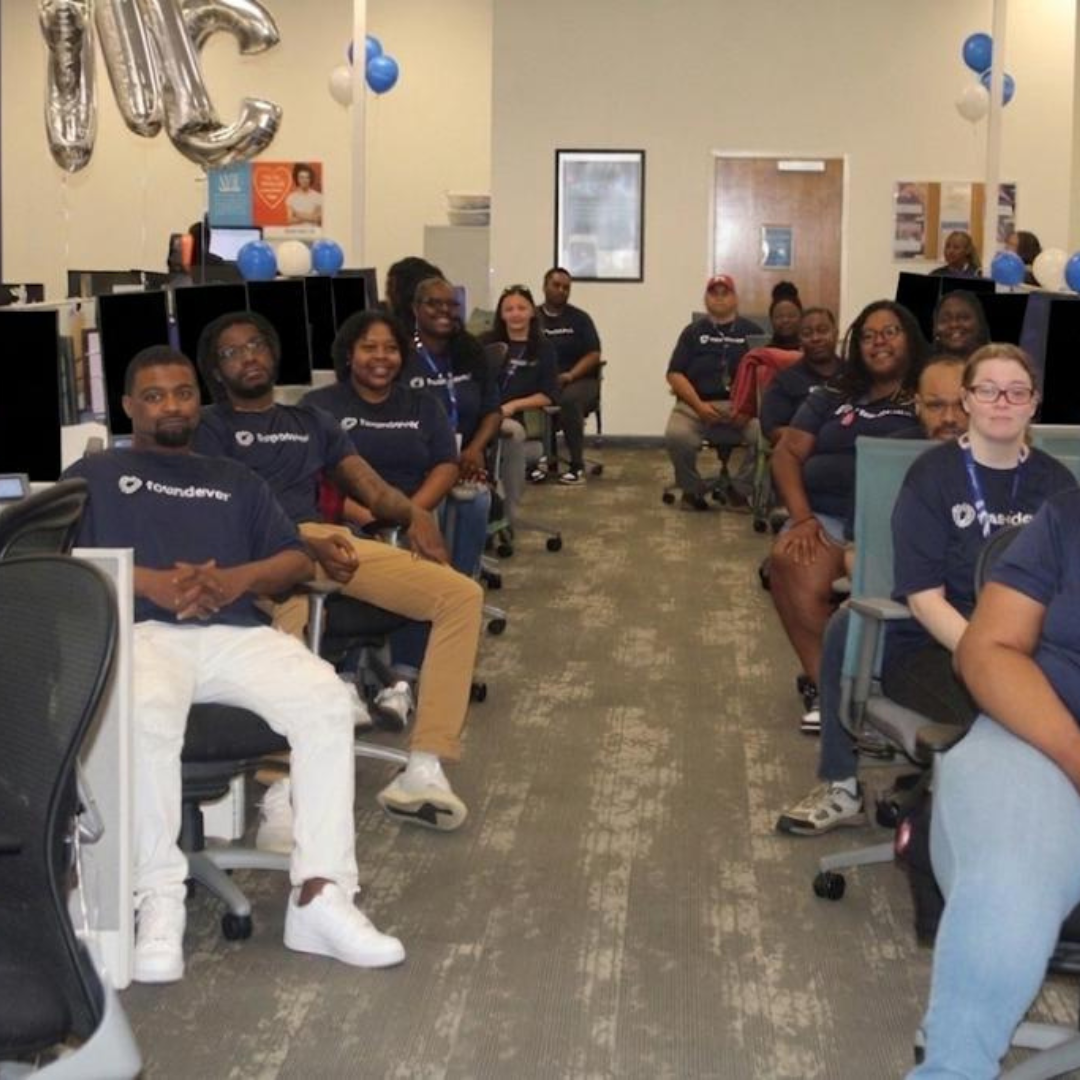 Two lines of individuals, chairs, and computers, they're all facing the camera and smiling but they are in an office space, at their desks, performing their job