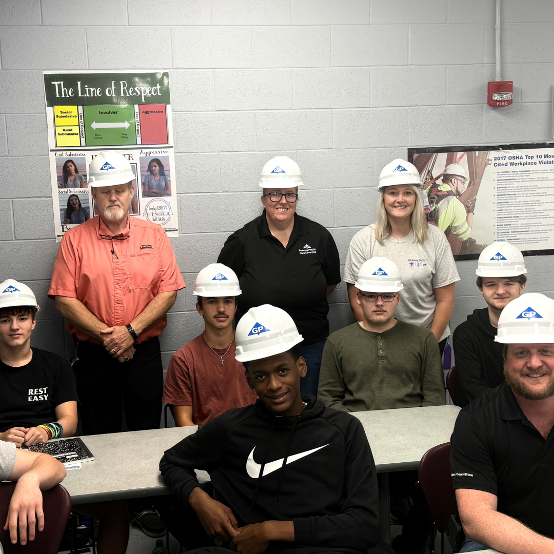 A group of people are sitting and standing around tables with hard hats smiling for the picture