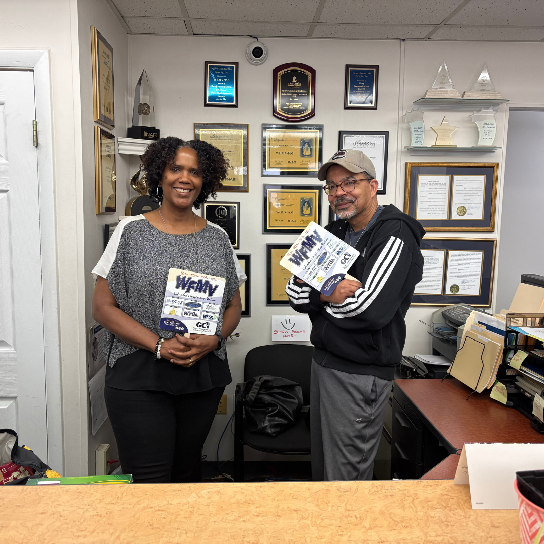Two individuals are standing behind a desk, both holding flyers on sticks