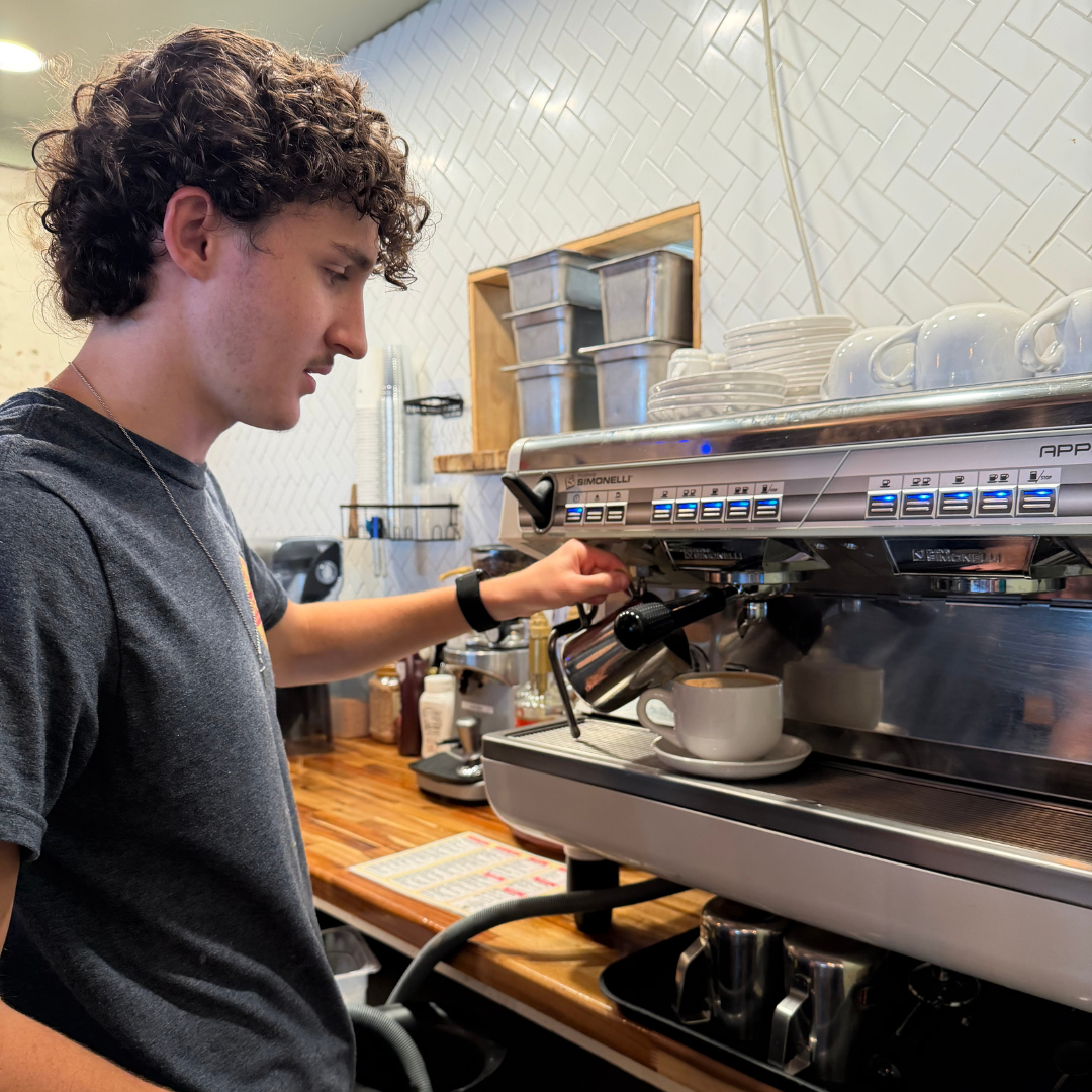 A man is standing in front of a coffee/ espresso machine, making a coffee 
