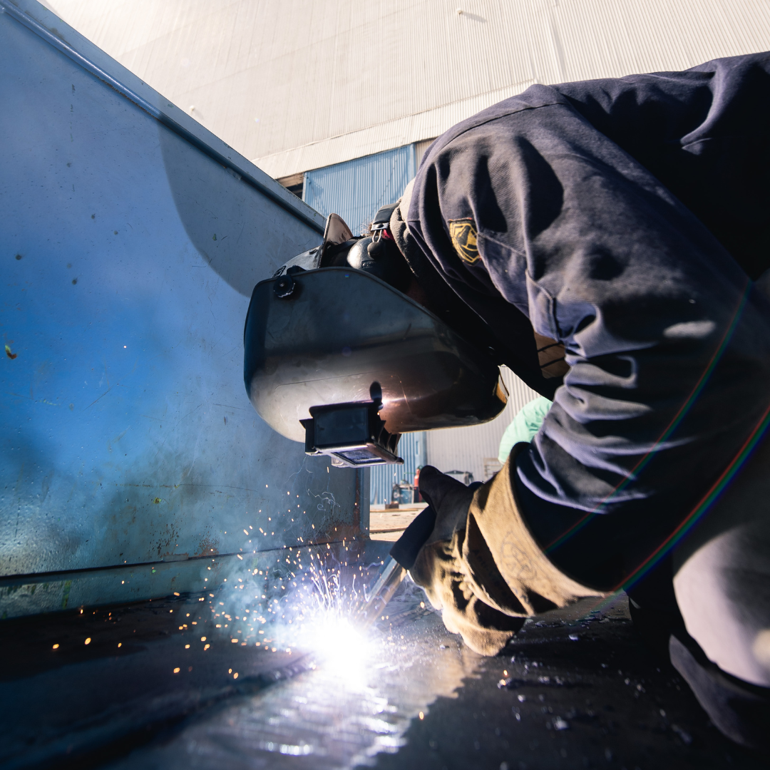 A man is leaned over a steel piece and he is using electric tools to weld the corners of the steel item. The man is in a protective visor helmet and sparks are flying from the fusion