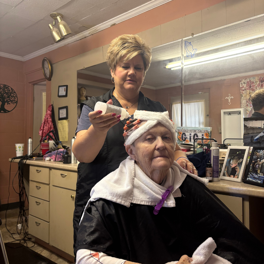 Two women are in a hair salon. One lady is behind the other, putting material in the women's hair. The older lady, is sitting in the chair, with a towel around her neck, and rollers in her hair