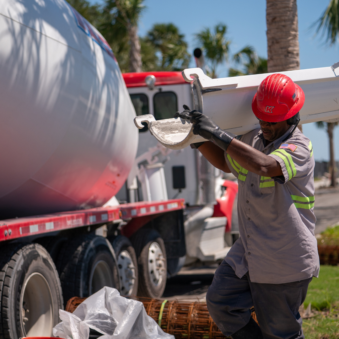 A man with a hard hat, holds a big beam, walking towards the left side of the picture. Behind him, a concrete mixing truck is to his right