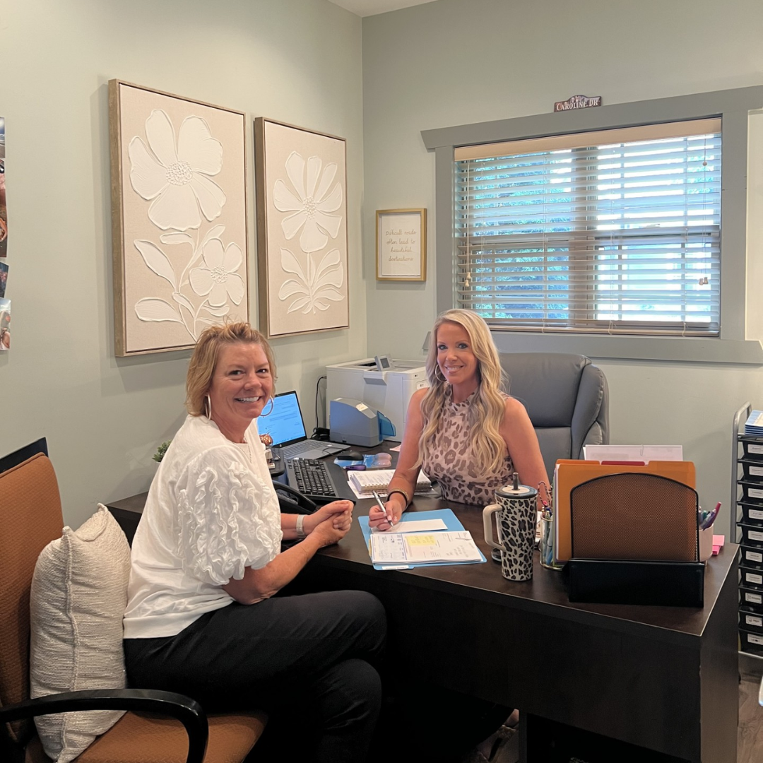 Two women sit on either side of the desk, conducting work. Both are facing the camera, smiling for the picture. 