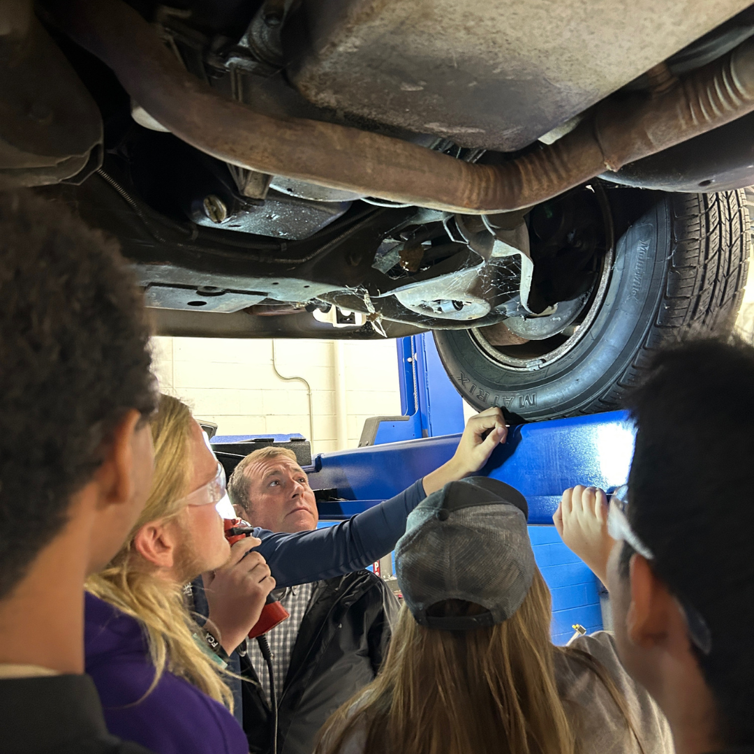 A group of students gather underneath a car with an instructor. They are observing the right tire of the car