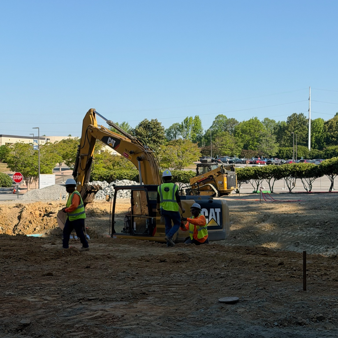 Several men in reflective vests work in a sandy area with an excavator