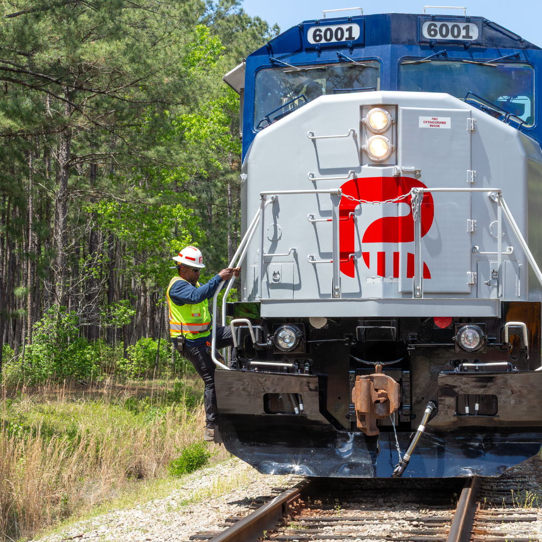 A train takes up most of the right side of the image, the train is near woods. A worker hangs off the back of the train