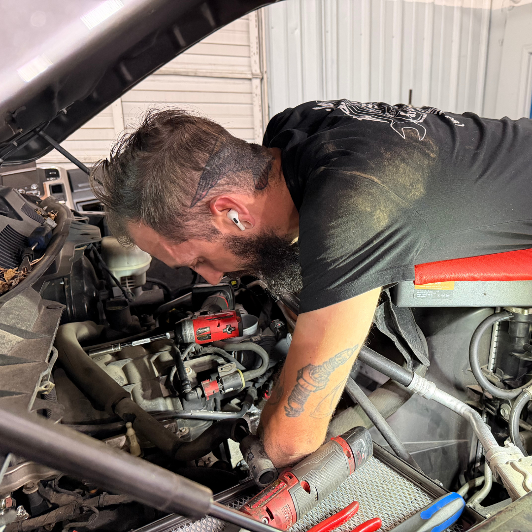A man with a uniformed shirt, is arms deep in the hood of the trunk, working to fix any issues