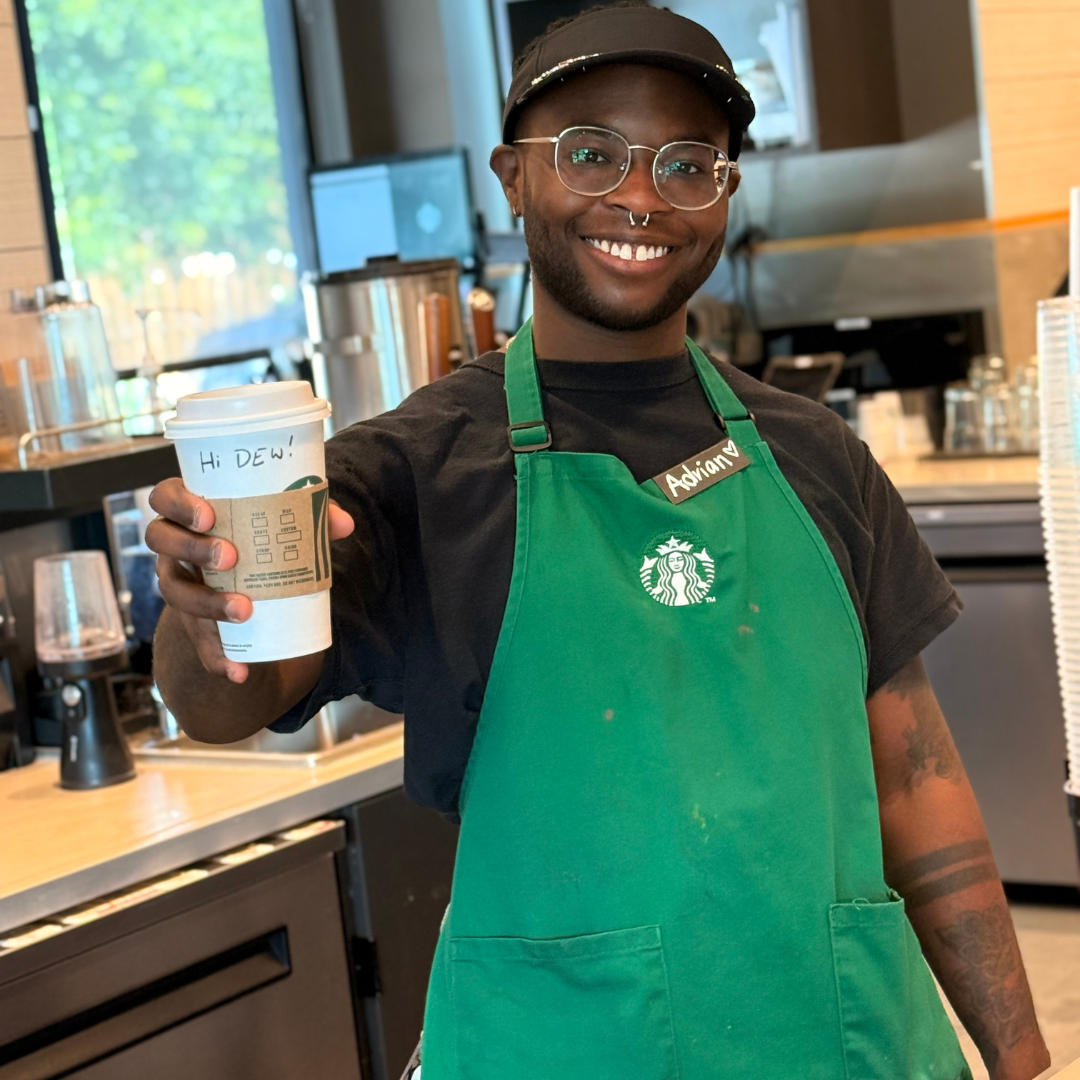 A male Starbucks worker is behind the counter, holding up a Starbucks cup that says 'Hi, DEW!' on it