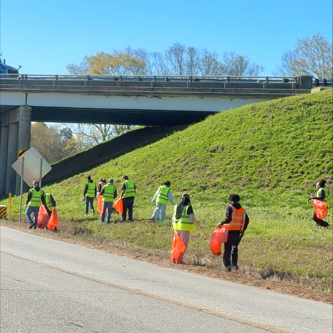Several individuals in a orange and yellow reflective vest, stand on the side of a road, picking up trash with a garbage picker