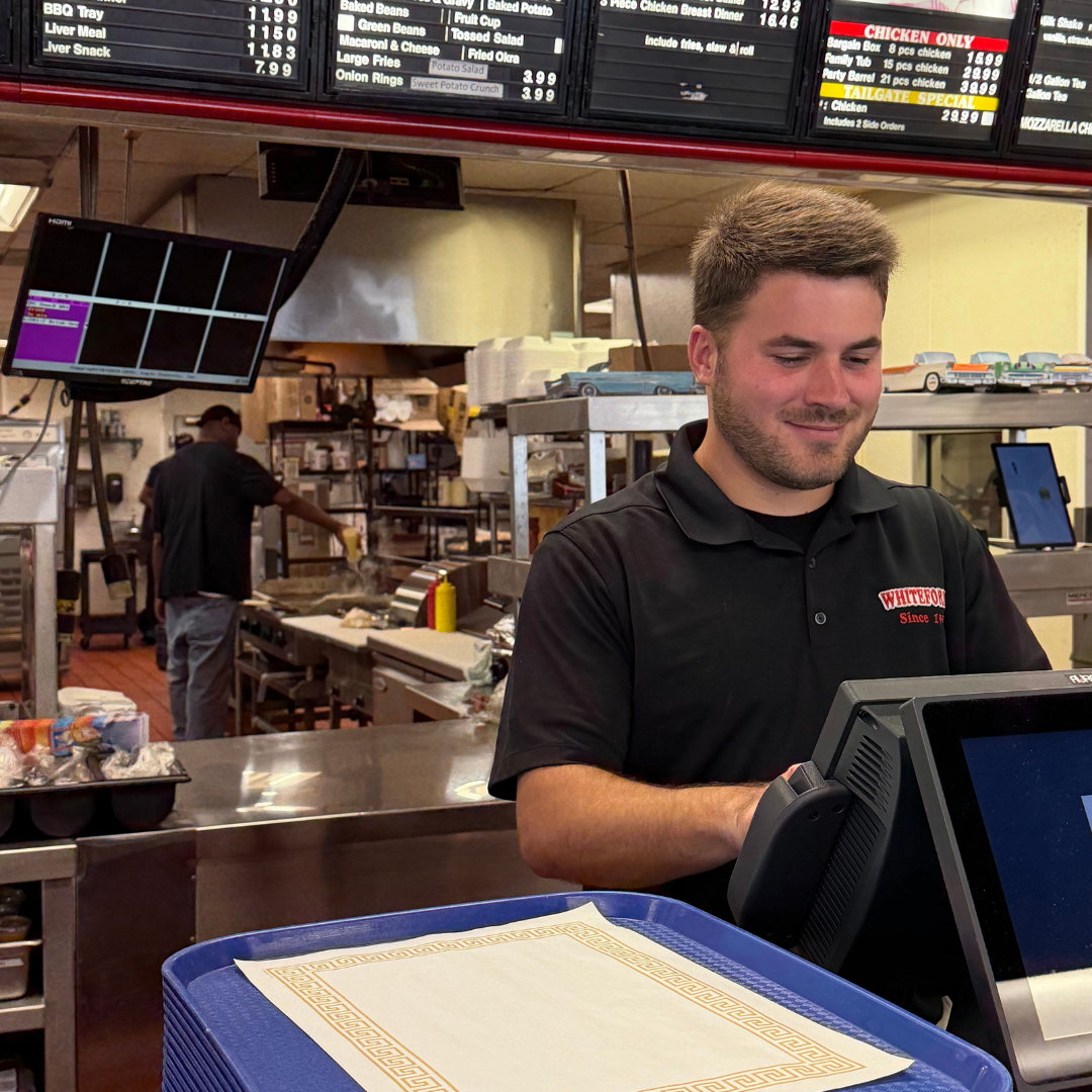 A man stands behind a counter at a fast-food restaurant. He is smiling, working the cash register. Behind him are boards displaying the menu items. 