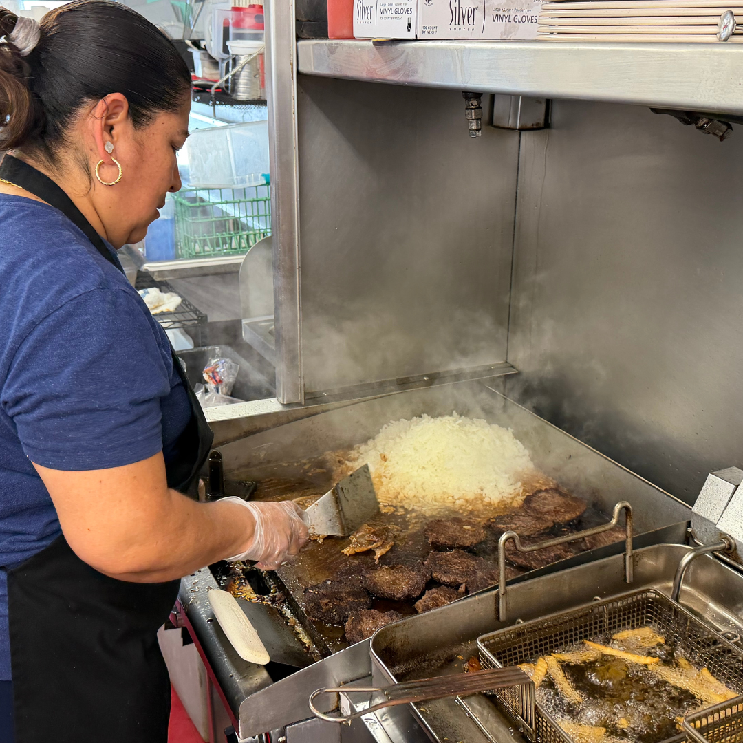 A woman with a black apron on stands in front of a grill with a spatula in her right hand. She is cooking white rice and  burgers on the grill. 