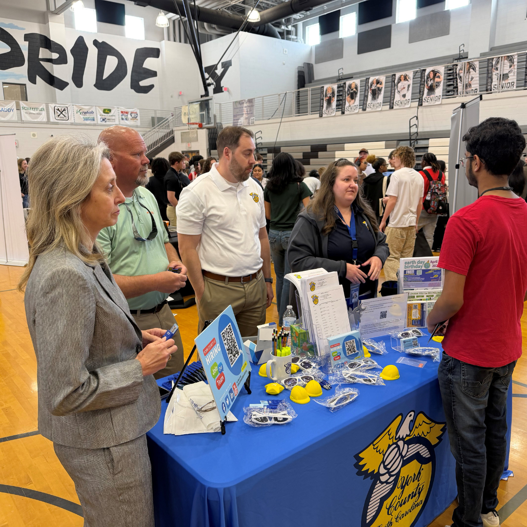 A group of people stand behind a blue booth in a gymnasium, they are speaking to a man on the other side of the booth. 