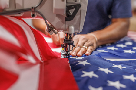 A hand with a gold ring is guiding fabric through a sewing machine. The fabric is used to make the American flag.