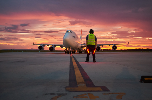 A person stands on a runway, holding two illuminated wands. Directly in front of him is an aircraft and it is poised under a pink and yellow sky.