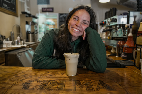 A young woman is smiling in front of an iced coffee in a plastic cup with a straw. She is leaned over a wooden bar.