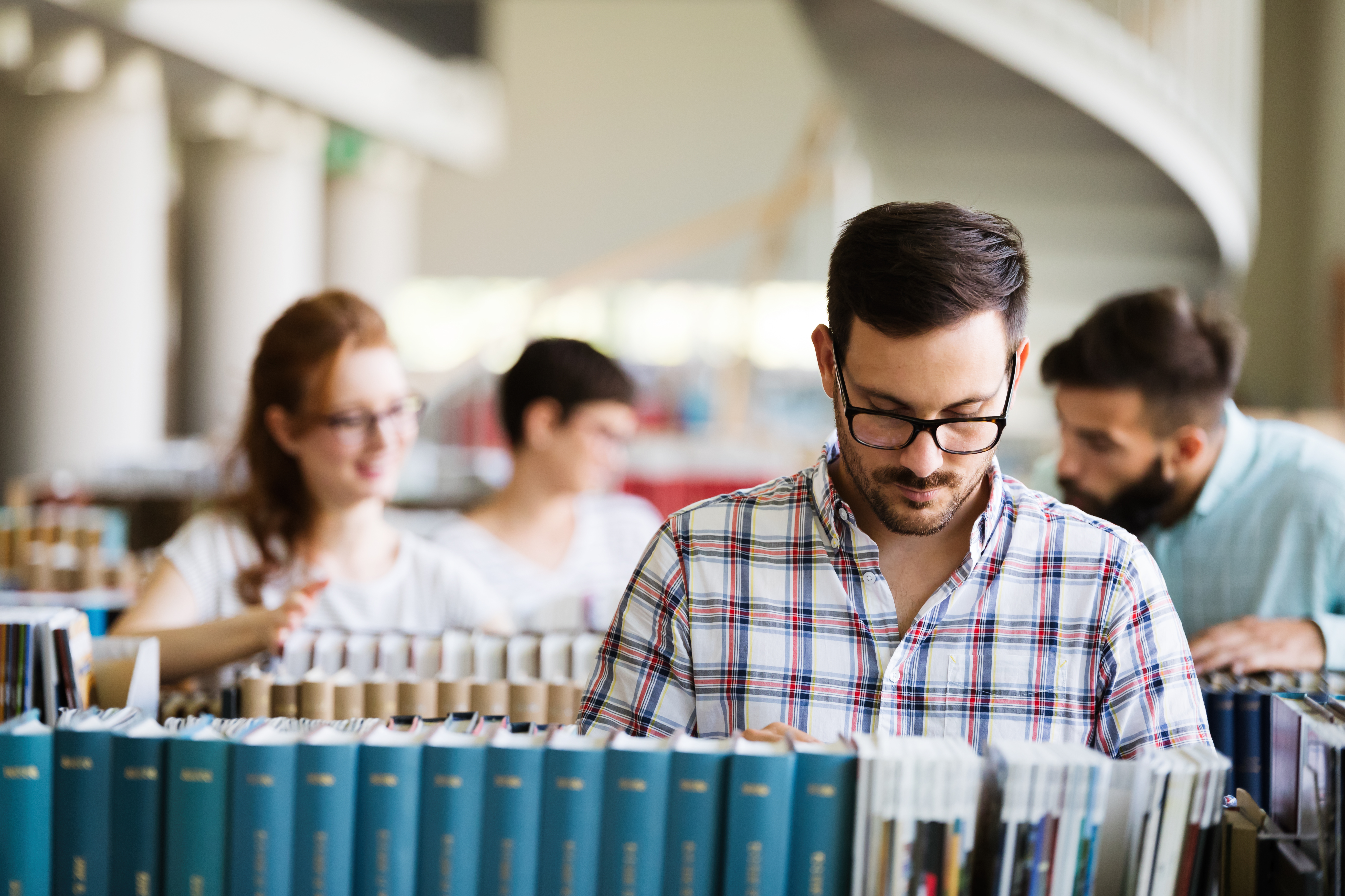 A man with glasses looks through a row of books. Three people look through books in the background behind him.