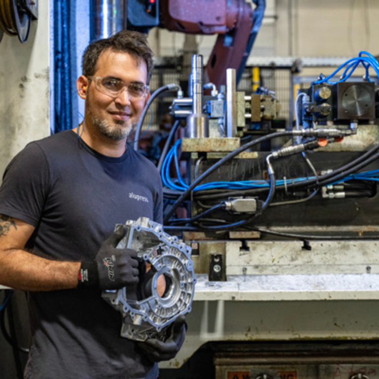 A man is holding a big steel item from his work, he is standing in front of machinery, smiling with protective glasses on 