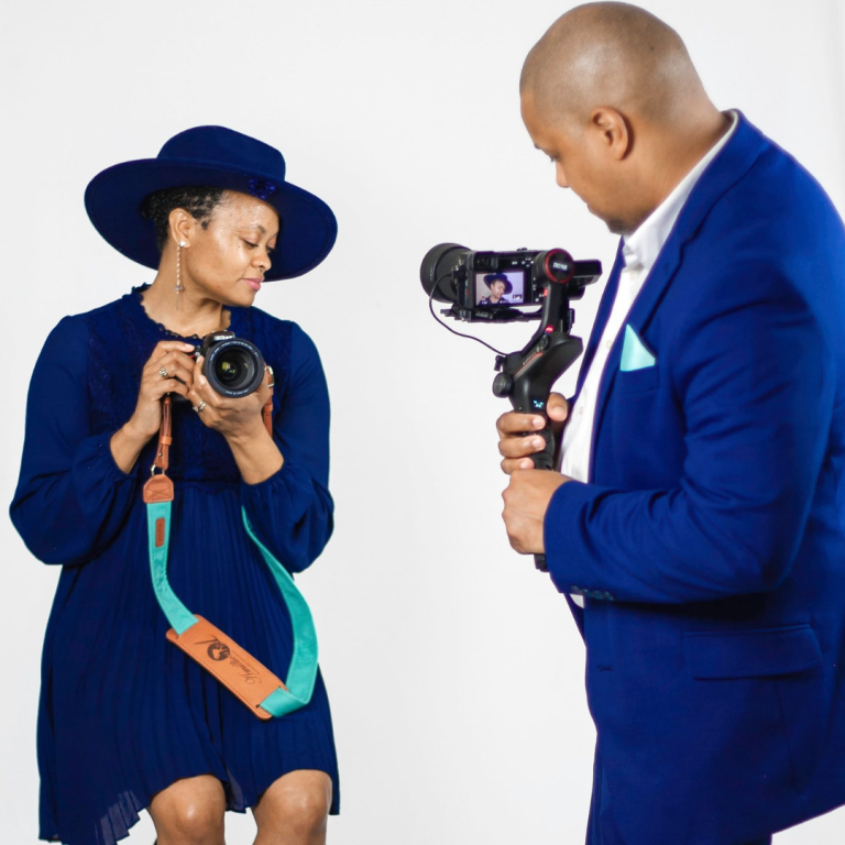 A man, nicely dressed in a suit, is taking a picture of a woman sitting on a stool, posing with a camera in hand and a big hat