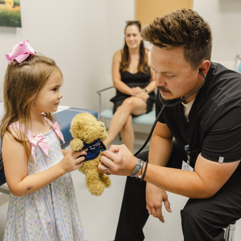 A male nurse is using his stethoscope to measure the heart beat of a stuffed bear a little girl is holding - her mom is in the background