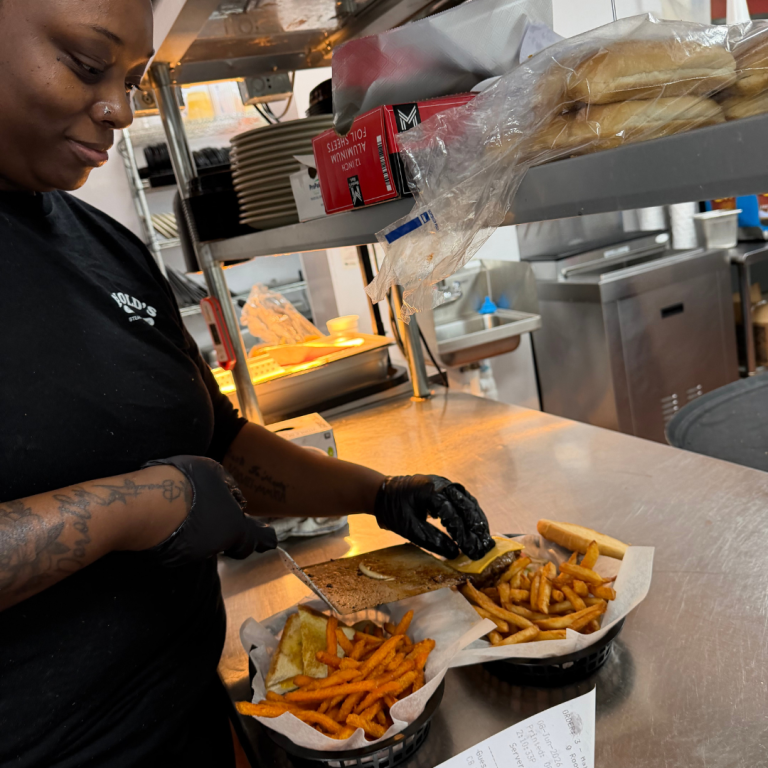 A woman is working in a restaurant, working on two meals in front of her in a basket to be served to customers, she is finishing one basket holding fries and a burger 