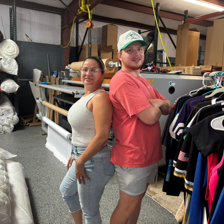 Two individuals, one woman, and one man, are standing back-to-back in a warehouse standing next to a rack of hung shirts