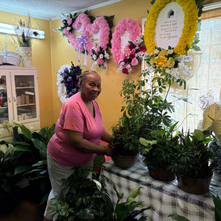 A woman is holding a potted plant, posing for a picture, surrounded by plants and a few flowers in a room