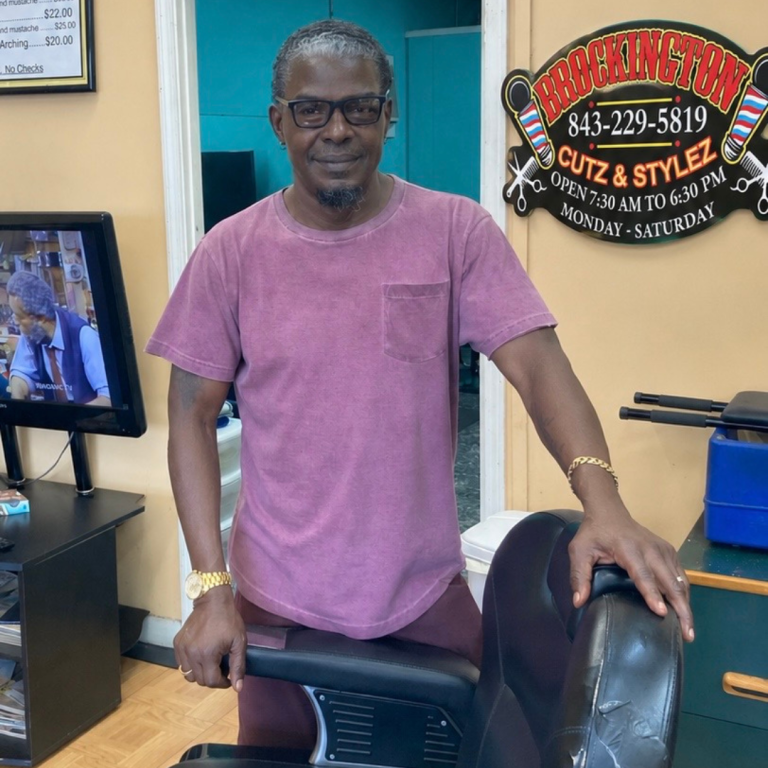 A man is standing in the building, Brockington Cutz & Stylez, standing in front of a barber chair, posing and smiling for the picture 