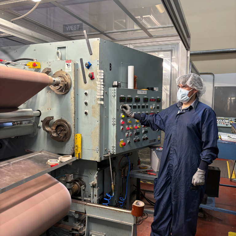 A person dressed in a head-to-toe zip up protective jumpsuit, and protective face mask, and hair net, is working next to machinery, pressing buttons and other items