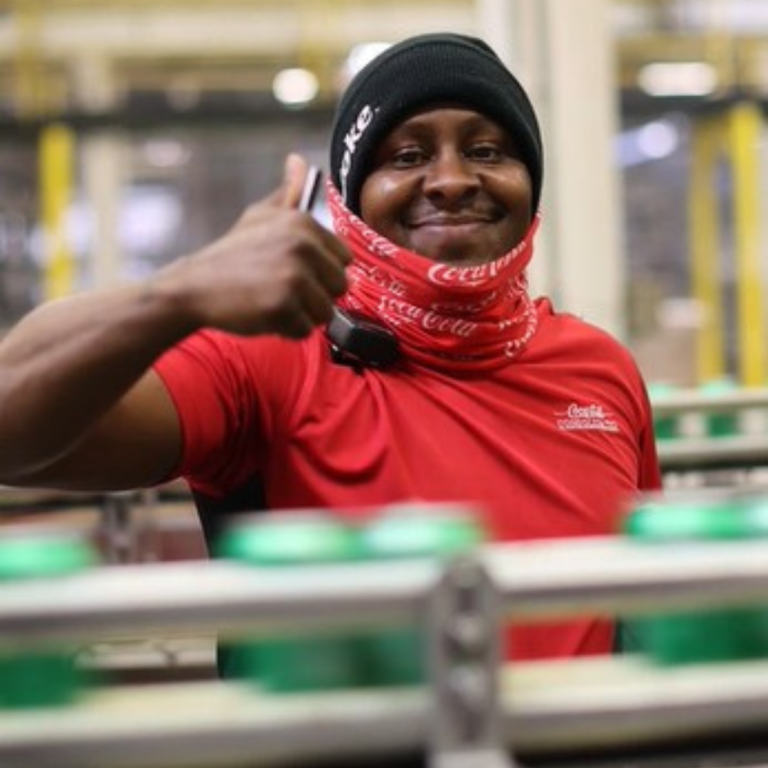 A man with a black beanie and a red, Coca-Cola face mask around his neck, is smiling and giving a thumbs-up to the camera