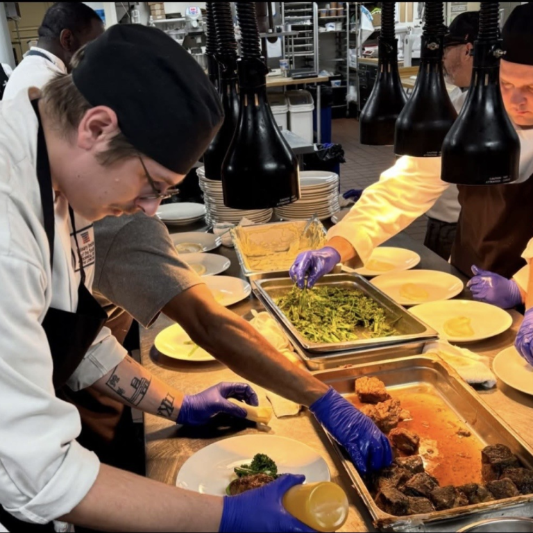 A group of chefs are around a table with plates of food on them, they are preparing and presenting the meals on their respective plates